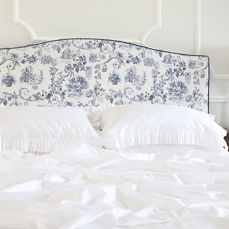 Elegant bed with a blue and white patterned headboard, white ruffled pillows, and crumpled white sheets, set against a paneled wall.