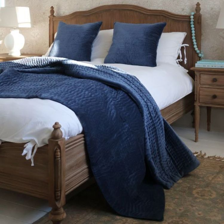 A cosy bedroom with a wooden bed, blue quilt and pillows, white lamps on wooden nightstands, and a decorative rug on the floor.
