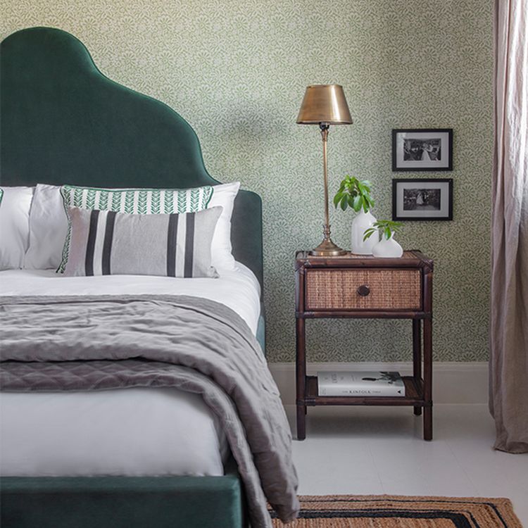 Cozy bedroom with green upholstered bed, patterned pillows, nightstand with lamp, plant, and framed photos on a patterned wallpapered wall.