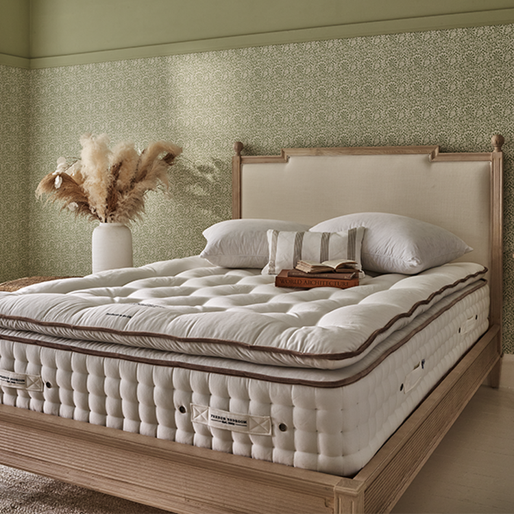 A cozy bedroom features a plush, white mattress on a wooden bed frame, with pillows and a book. A vase with pampas grass decorates the room.