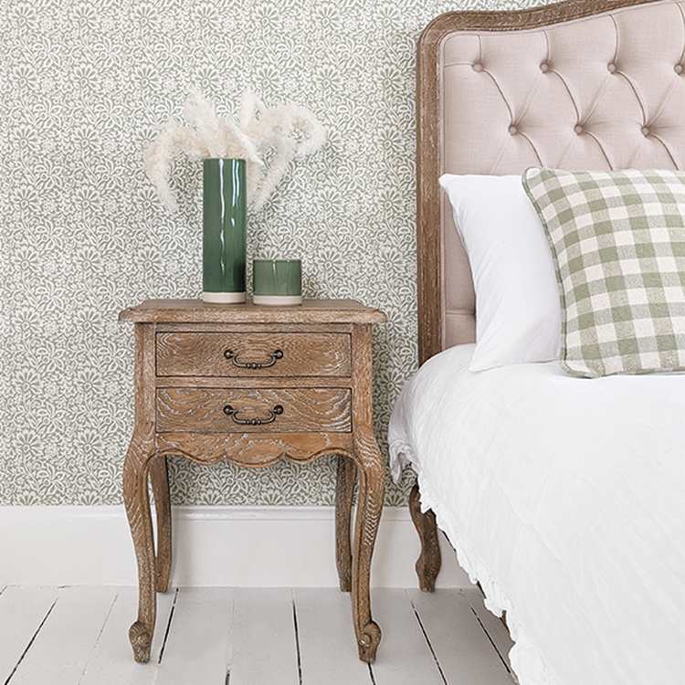 Elegant bedroom with a wooden nightstand, tufted headboard, patterned wallpaper, and decorative vases with pampas grass.