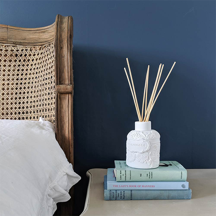 A white diffuser with reeds on a stack of books beside a wicker chair against a dark blue wall.