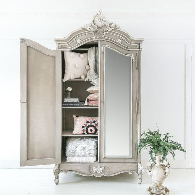Elegant vintage wardrobe with open doors, displaying neatly stacked pillows and a folded blanket. A decorative plant stands nearby.