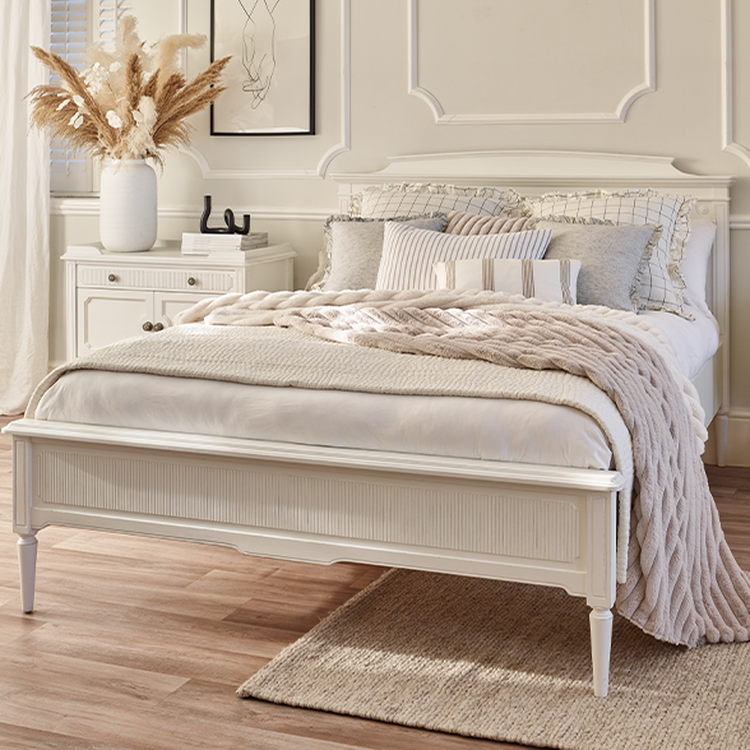 A cozy bedroom with a white bed, beige and white bedding, a knitted throw, a nightstand, and a vase of dried flowers on a wooden floor.
