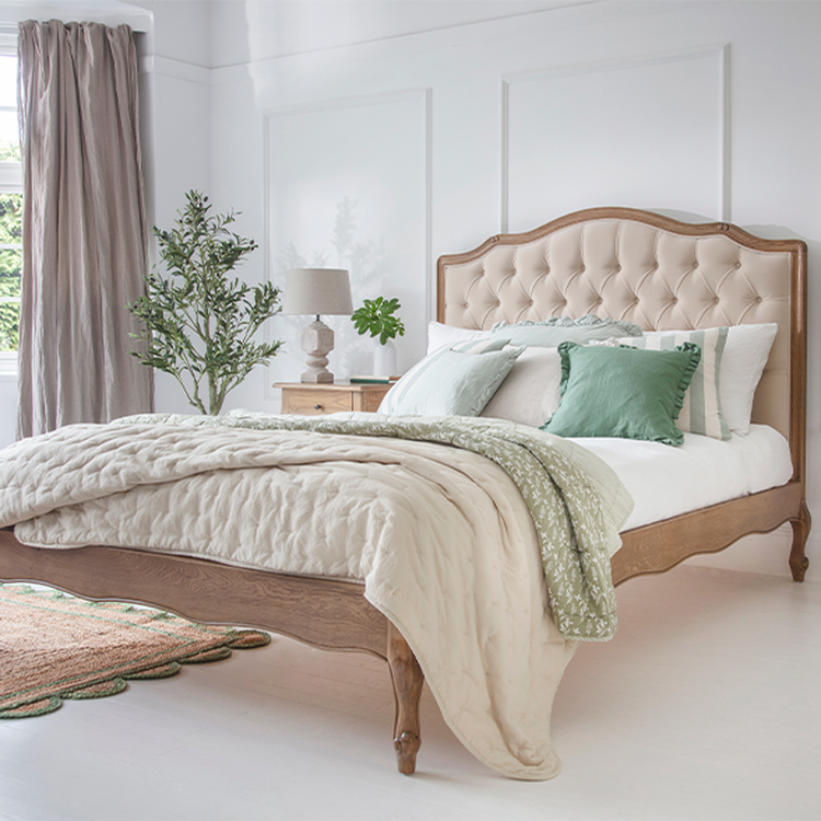 Elegant bedroom with a wooden bed, tufted headboard, green and white bedding, bedside table with lamp, potted plant, and window with curtains.
