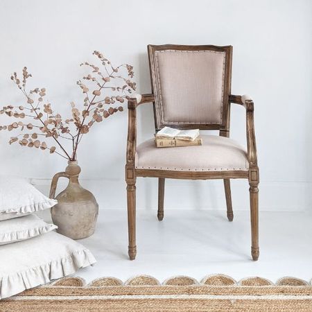 Elegant wooden chair with beige upholstery beside a rustic vase with dried branches, stacked pillows, and a woven rug on a light floor.