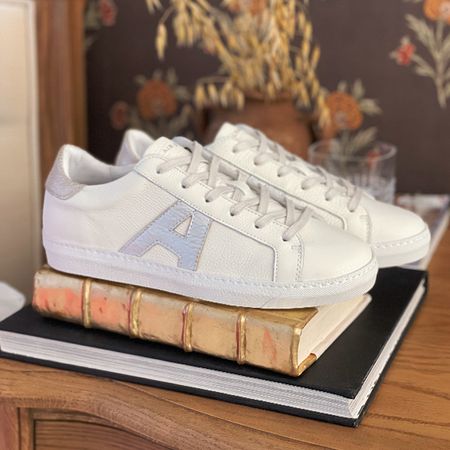 White sneakers with a silver "A" logo on the side, placed on stacked books with a floral wallpaper background.