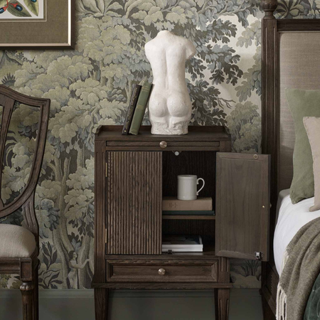Bedroom scene with a wooden nightstand, a torso sculpture on top, books, a mug inside, and floral wallpaper in the background.