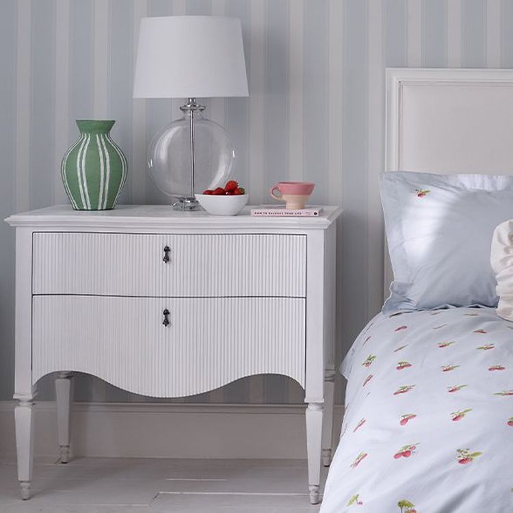 A white bedside table with a green vase, clear lamp, and bowl of strawberries. A bed with patterned sheets is beside it, against striped wallpaper.