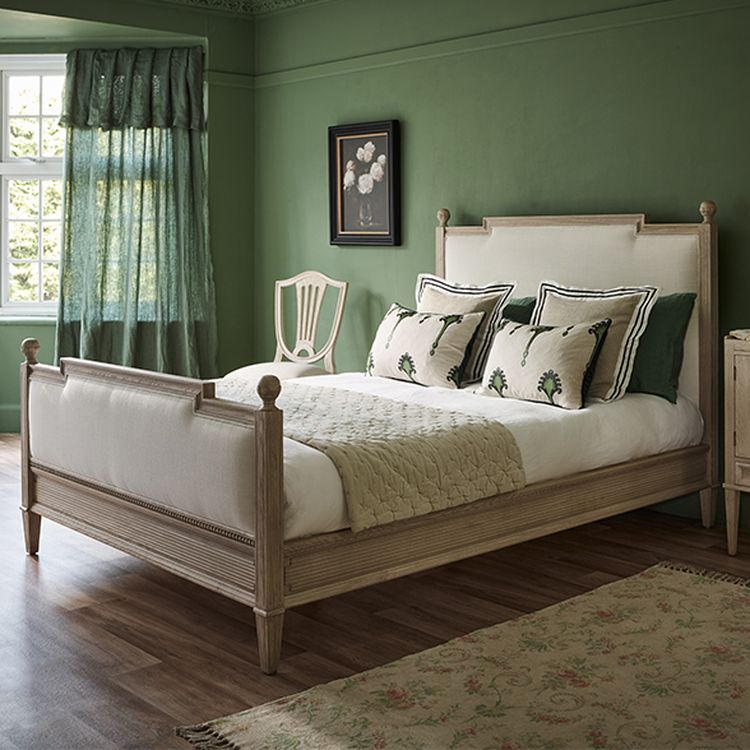 Elegant bedroom with a wooden bed, green walls, floral pillows, and a decorative chair. Sunlight filters through sheer curtains.