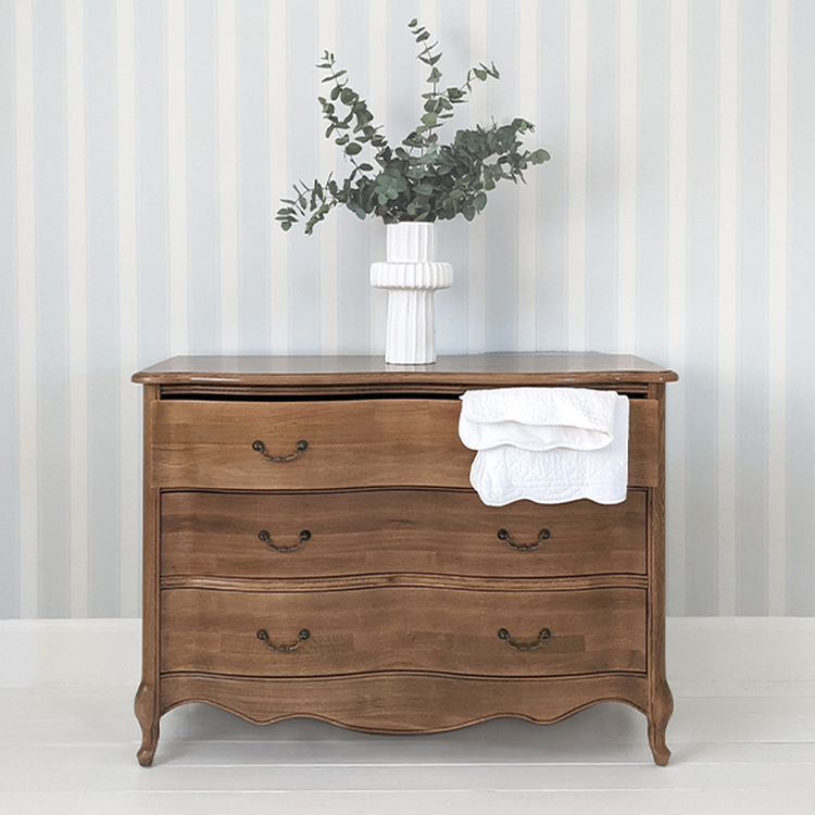 Wooden dresser with three drawers, a white vase holding eucalyptus branches on top, and a folded white cloth hanging from a drawer, set against striped wallpaper.