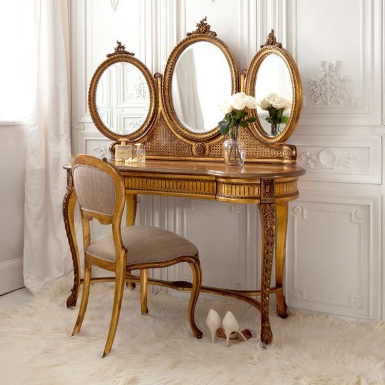 Elegant vanity table with ornate gold details, three mirrors, a cushioned chair, white roses, and a pair of white heels on a plush rug.