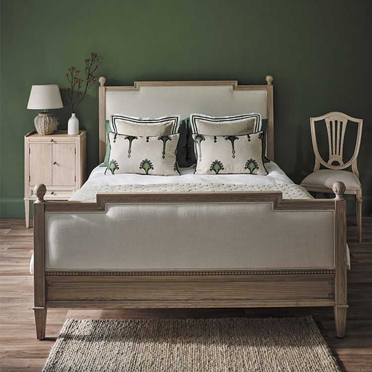 Elegant bedroom with a wooden bed, decorative pillows, a nightstand with a lamp, and a chair against a green wall. Rug on wooden floor.