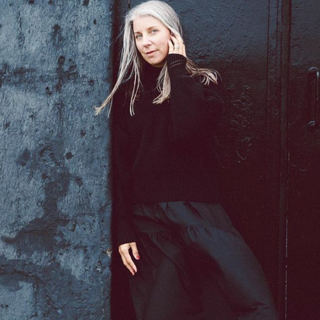 Woman with long gray hair in a black outfit leans against a textured wall, looking at the camera with a calm expression.