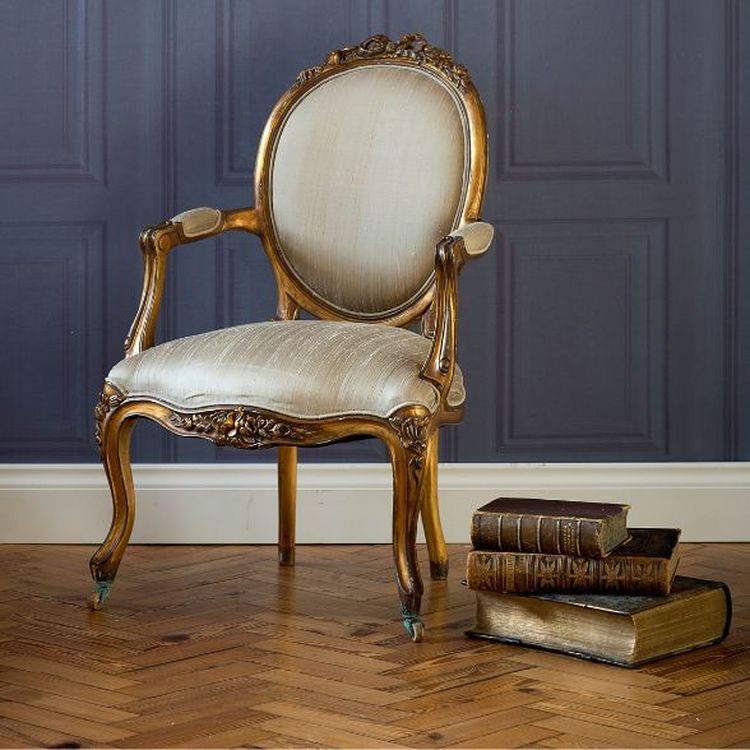 Elegant antique chair with ornate wooden frame and light upholstery beside a stack of vintage books on a herringbone wood floor.