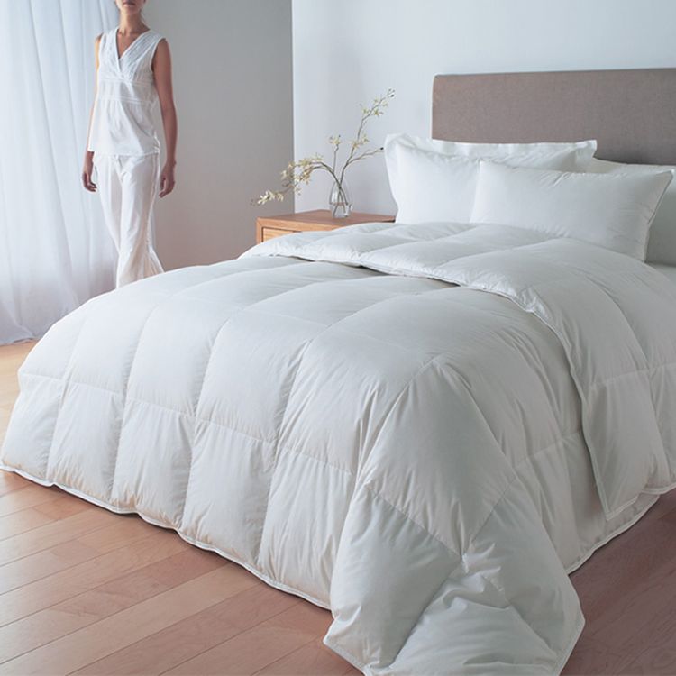 A neatly made bed with a white comforter and pillows in a bright room. A person in white pajamas stands nearby.