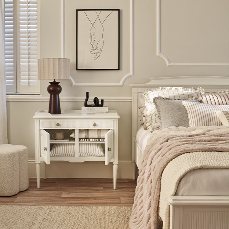 Cozy bedroom with a white nightstand, lamp, and framed artwork. Bed with striped pillows and a soft pink blanket. Neutral tones throughout.