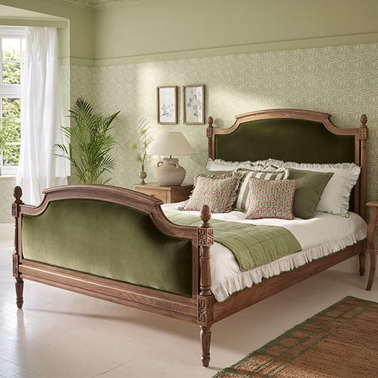 Elegant bedroom with a wooden bed frame, green upholstered headboard, matching pillows, and plants by a window. Soft green walls and patterned decor.