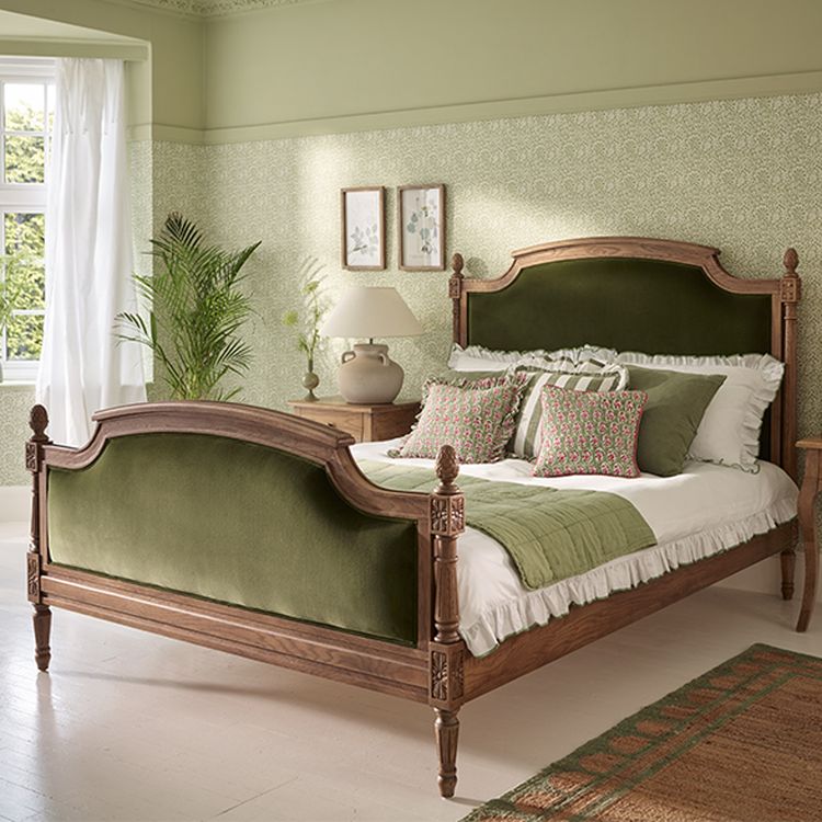 Elegant bedroom with a wooden bed frame, green upholstered headboard, matching pillows, and plants by a window. Soft green walls and patterned decor.