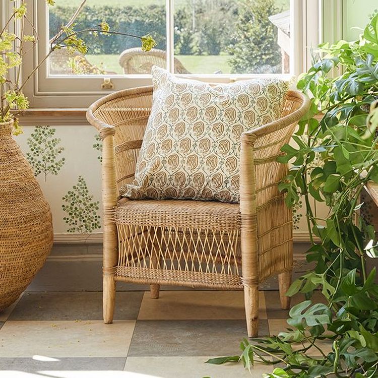 Wicker chair with a patterned cushion in a sunlit room, surrounded by green plants and a large window overlooking a garden.