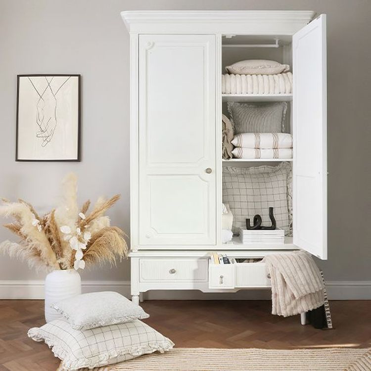 White wardrobe with open door revealing neatly folded linens. Nearby are a vase with pampas grass, a framed line drawing, and checkered pillows.
