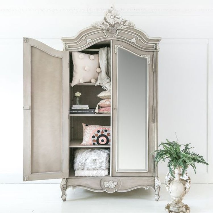Ornate vintage wardrobe with open door, revealing shelves of decorative pillows and throws, next to a potted fern on a white floor.