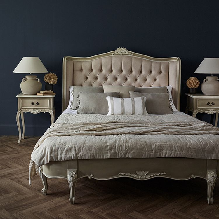 Elegant bedroom with a tufted headboard, beige bedding, two vintage nightstands, and lamps against a dark wall on herringbone wood flooring.