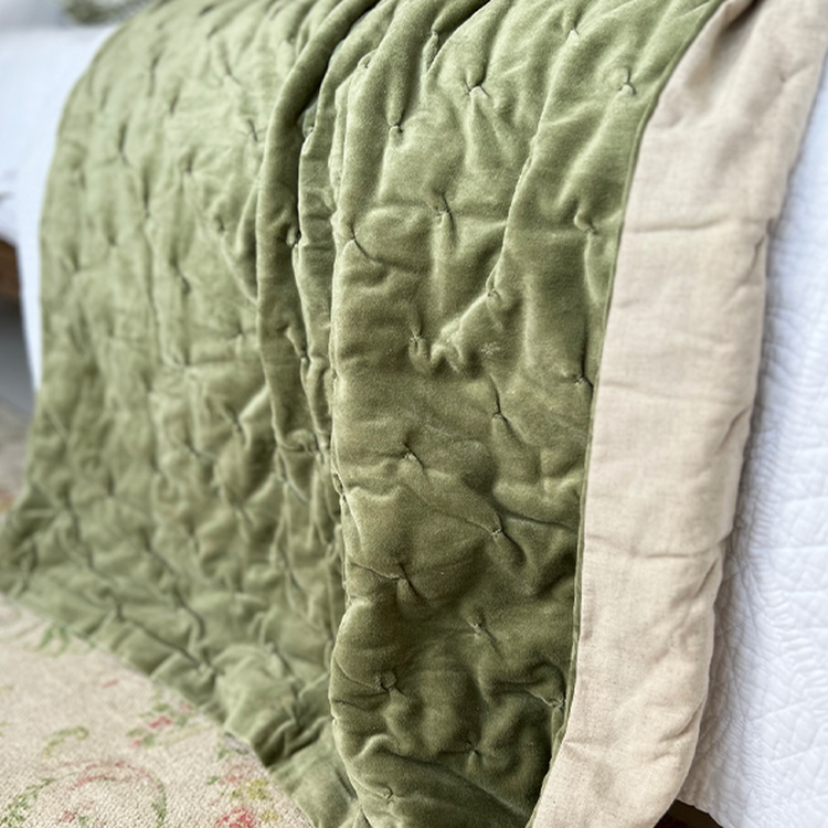 A soft, green quilt with a tufted pattern drapes over the edge of a bed, revealing a beige underside.