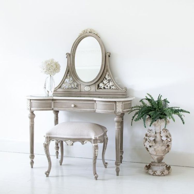 Elegant vintage vanity set with ornate mirror, floral carvings, cushioned stool, and a decorative vase with greenery against a white wall.