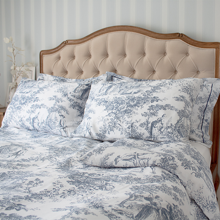 Elegant bed with a tufted headboard, adorned with blue and white patterned bedding featuring pastoral scenes, in a softly lit bedroom.