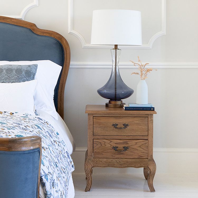 Bedroom scene with a wooden nightstand, table lamp, and vase. A bed with a blue headboard and floral bedding is partially visible.