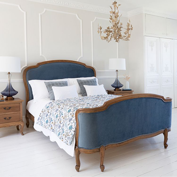 Elegant bedroom with a blue velvet bed, floral bedding, ornate chandelier, and classic wooden furniture against white walls and flooring.