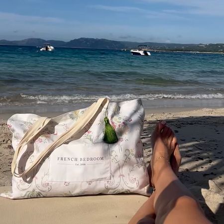 A floral beach bag on sandy shore, with a person's legs, overlooking a calm sea and distant boats under a clear sky.