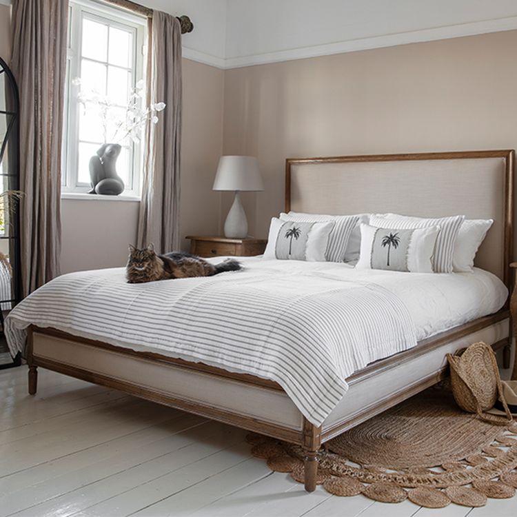 Cozy bedroom with a wooden bed, white bedding, decorative pillows, a cat lounging, and a window with curtains. Neutral tones and natural light.