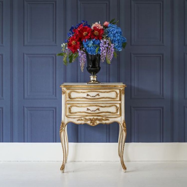 Elegant ornate table with a black vase holding vibrant red, pink, and blue flowers against a textured blue paneled wall.