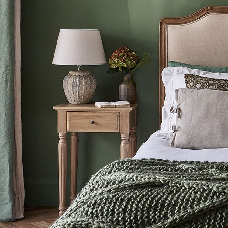 Cozy bedroom with a wooden nightstand, lamp, and vase beside a bed with green and white bedding and a knitted throw. Green wall backdrop.