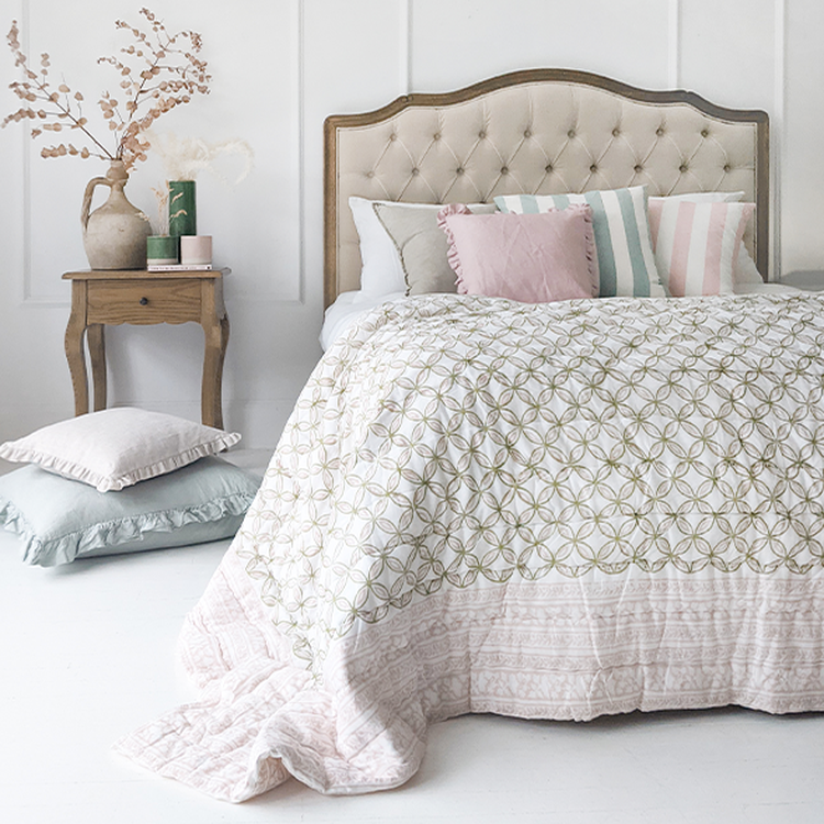 Elegant bedroom with a tufted headboard, patterned quilt, pastel pillows, and a wooden side table with decorative vases and dried flowers.