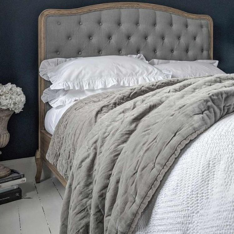 Cozy bedroom with a tufted headboard, fluffy pillows, a gray quilted blanket, and a vase of white flowers on a stack of books.