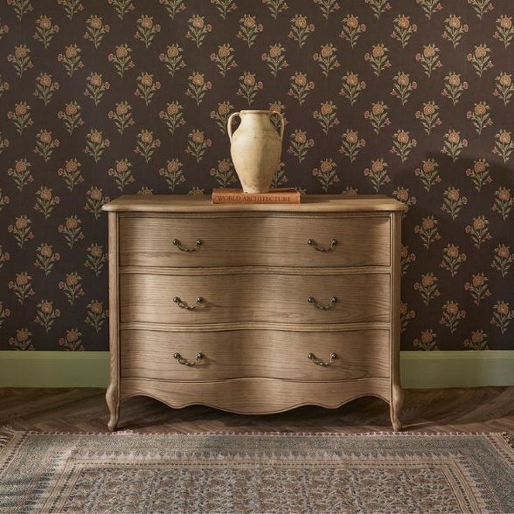 Wooden dresser with curved drawers, a ceramic vase, and a book on top, set against floral wallpaper and a patterned rug.