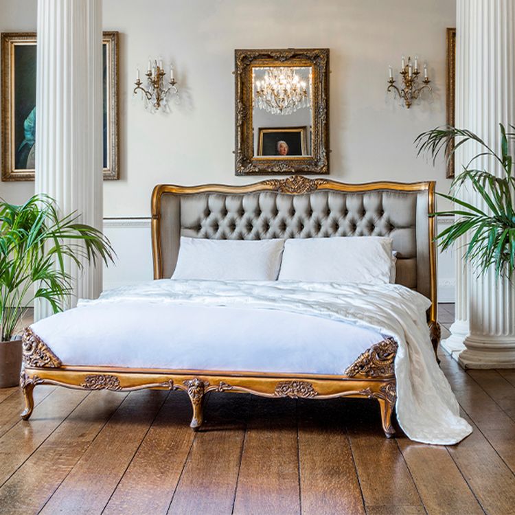 Elegant bedroom with a tufted headboard bed, ornate gold frame, white bedding, wooden floor, chandeliers, framed paintings, and potted plants.