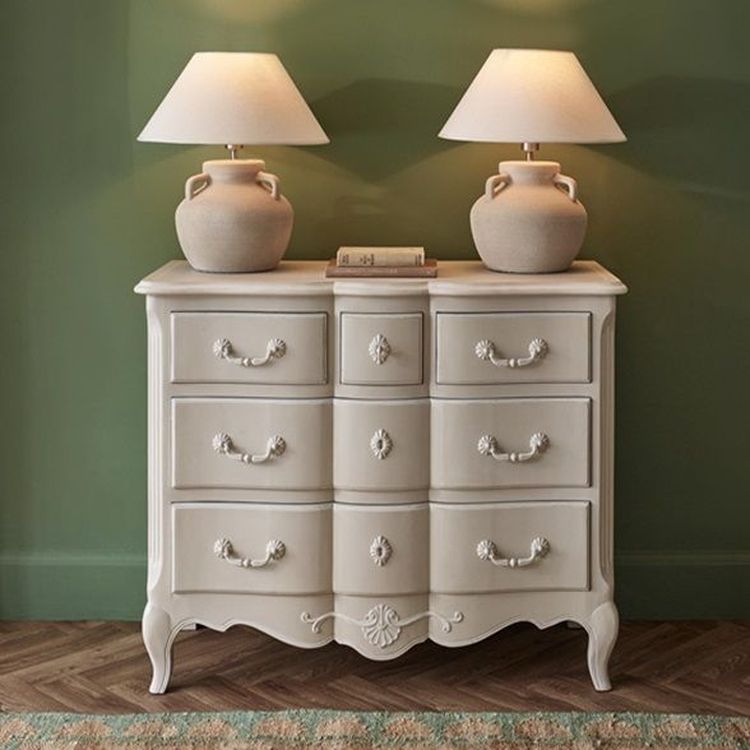 Cream-colored dresser with ornate handles, topped with two matching lamps and a book, set against a green wall and wooden floor.