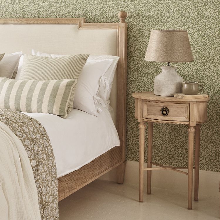 Cozy bedroom with a wooden bed, patterned green wallpaper, striped pillows, and a small wooden nightstand with a lamp and mug.