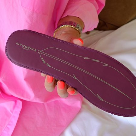 A person in a pink outfit holds a purple shoe insole with a feather design and "Air & Grace London" text, showcasing bright coral nail polish.
