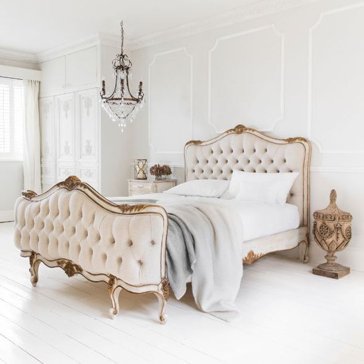 Elegant bedroom with a vintage-style bed, tufted headboard, ornate chandelier, and white walls. Light floods through a window with sheer curtains.
