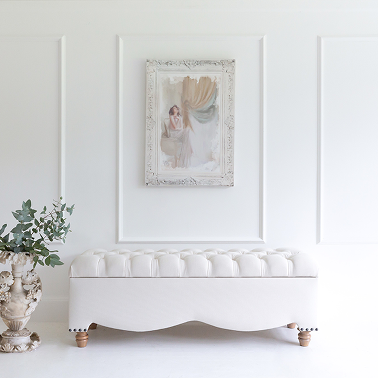 Elegant room with a white tufted bench, ornate vase with greenery, and a framed painting on a paneled wall.