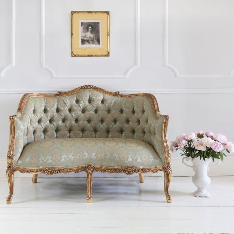 Elegant vintage sofa with ornate woodwork, tufted upholstery, and floral pattern. Framed portrait above; vase of pink flowers on the floor.