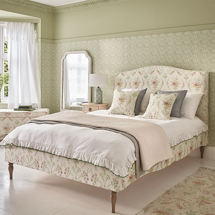 A cozy bedroom with a floral-patterned bed, matching pillows, a soft beige throw, a bedside lamp, a mirror, and light green walls.