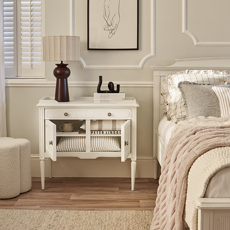 A cozy bedroom corner with a white nightstand, lamp, and abstract wall art. Soft pillows and a beige throw blanket adorn the bed.