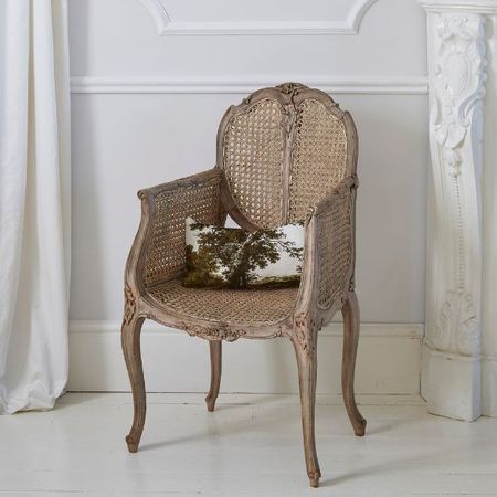 Ornate wooden chair with cane back and seat, featuring a decorative pillow with a tree design, set against a white wall and draped curtain.
