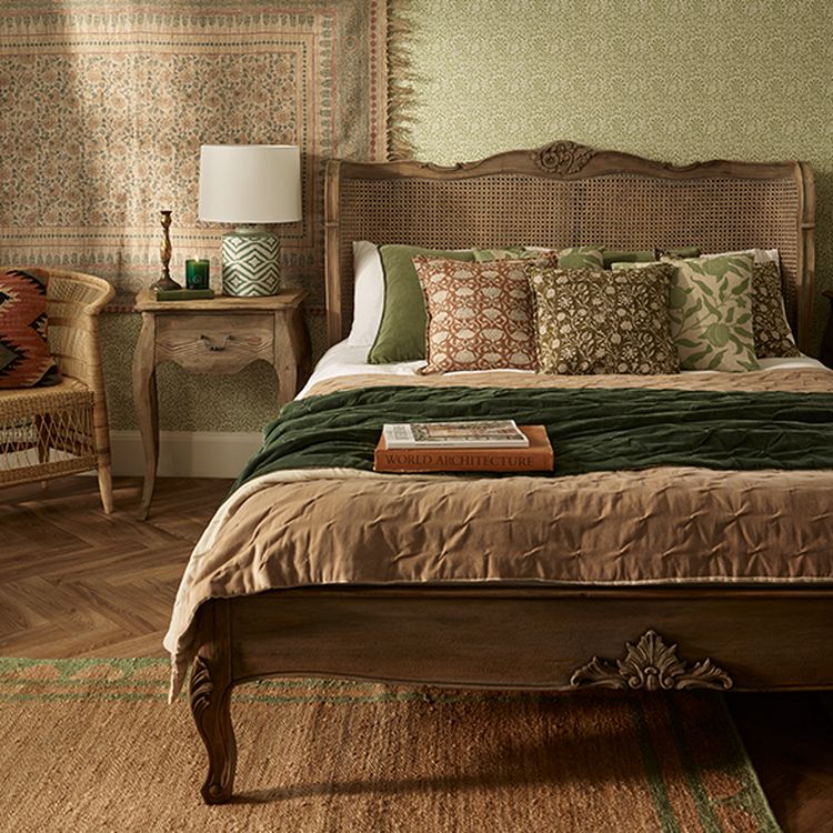 Cozy bedroom with a vintage wooden bed, patterned pillows, green and beige bedding, a woven chair, and a side table with a lamp and book.